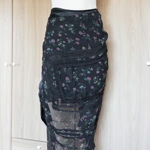 Peut inclure: Jupe noire &agrave; imprim&eacute; floral avec une superposition transparente et une bordure en dentelle. La jupe pr&eacute;sente une taille haute et un design asym&eacute;trique. Le motif floral comprend des fleurs roses et violettes.