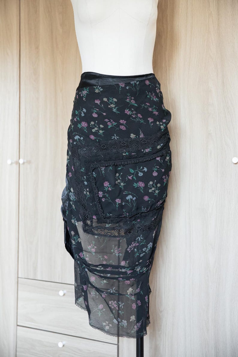 Peut inclure: Jupe noire &agrave; imprim&eacute; floral avec une superposition transparente et une bordure en dentelle. La jupe pr&eacute;sente une taille haute et un design asym&eacute;trique. Le motif floral comprend des fleurs roses et violettes.