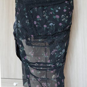 Peut inclure: Jupe noire &agrave; imprim&eacute; floral avec superposition transparente. Le motif floral pr&eacute;sente des fleurs roses, violettes et bleues. Des bordures en dentelle noire ornent les bords. La jupe est faite d'un tissu l&eacute;ger et fluide.