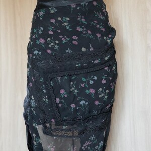 Peut inclure: Jupe noire &agrave; imprim&eacute; floral avec une d&eacute;licate bordure en dentelle. La jupe pr&eacute;sente un motif de petites fleurs roses et violettes sur un fond sombre. La jupe est faite d'un tissu transparent.