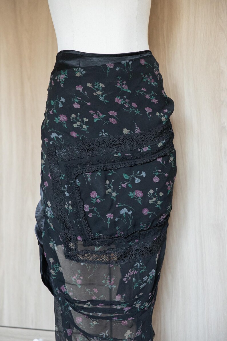 Peut inclure: Jupe noire &agrave; imprim&eacute; floral avec une d&eacute;licate bordure en dentelle. La jupe pr&eacute;sente un motif de petites fleurs roses et violettes sur un fond sombre. La jupe est faite d'un tissu transparent.
