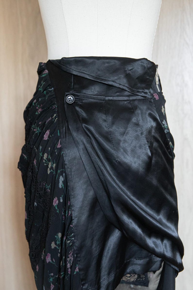 Peut inclure: Jupe noire au design unique. La jupe pr&eacute;sente un rev&ecirc;tement en satin qui recouvre un tissu &agrave; motifs floraux. Une fermeture &agrave; bouton et &agrave; glissi&egrave;re est visible. Le tissu floral a une bordure en dentelle.