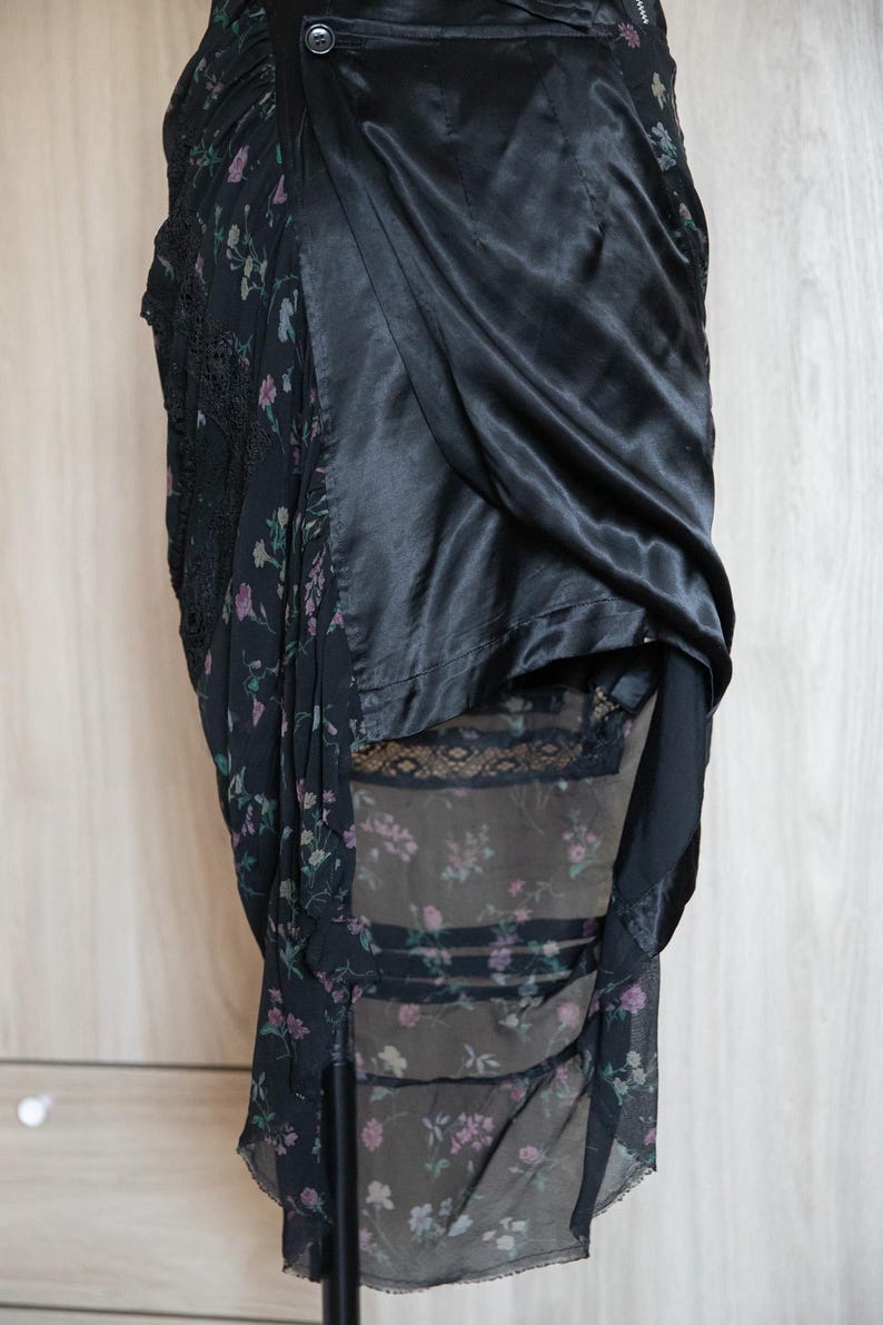 Peut inclure: Jupe noire avec imprim&eacute; floral et d&eacute;tails en dentelle. La jupe pr&eacute;sente un rev&ecirc;tement en satin drap&eacute; et une sous-couche florale transparente. Le motif floral comprend des fleurs roses et violettes sur fond sombre. Fermeture boutonn&eacute;e.