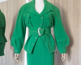 May include: A vibrant green jacket and skirt set featuring a collared jacket with long puffed sleeves, button closures, two chest pockets, and a matching belt with a silver-toned buckle. The skirt is a simple straight-cut design in the same green.