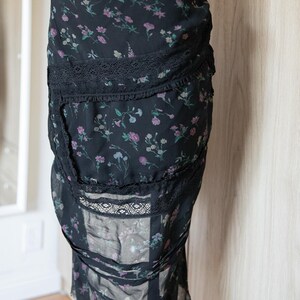 Peut inclure: Jupe noire &agrave; imprim&eacute; floral avec bordure en dentelle. La jupe pr&eacute;sente un motif d&eacute;licat de fleurs roses, violettes et vertes sur fond noir. La jupe est faite d'un tissu l&eacute;ger et a une silhouette fluide.