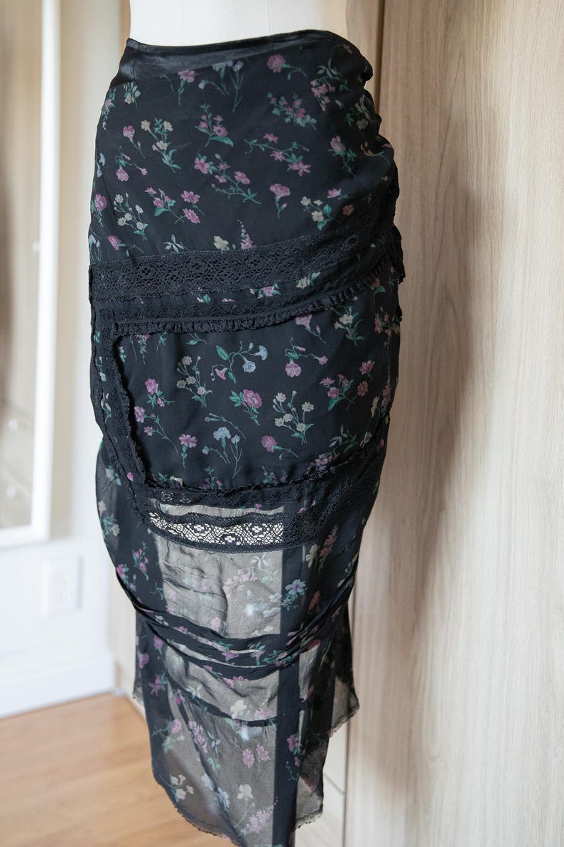 Peut inclure: Jupe noire &agrave; imprim&eacute; floral avec bordure en dentelle. La jupe pr&eacute;sente un motif d&eacute;licat de fleurs roses, violettes et vertes sur fond noir. La jupe est faite d'un tissu l&eacute;ger et a une silhouette fluide.