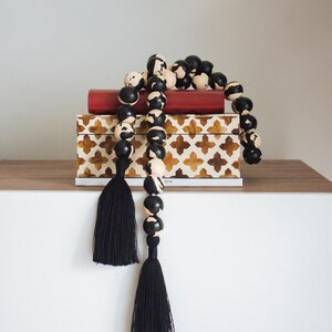 May include: A wooden beaded garland with black and cream accents and two black tassels. The garland is draped over a red book and a decorative box with a brown and white geometric pattern.