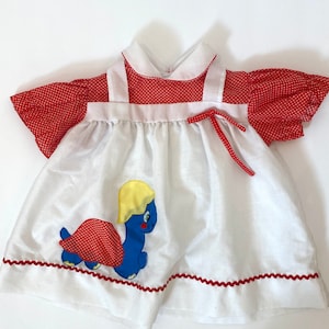 May include: A white baby dress with red polka dot sleeves and a red and white polka dot turtle applique. The dress has a white collar and a red ribbon tie.