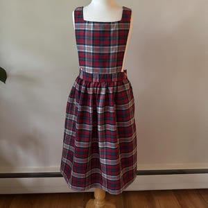 Puede incluir: Un vestido de peto a cuadros en rojo, azul marino y blanco, exhibido en un maniquí. El vestido presenta un escote cuadrado, un corpiño ajustado y una falda fruncida. La tela tiene un patrón de tartán clásico.