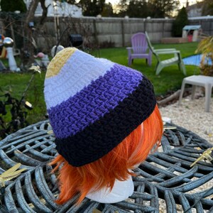 May include: A crocheted beanie hat with yellow, white, purple, and black stripes. The hat is on a mannequin head with orange hair.