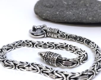 May include: A silver-toned chain bracelet featuring a complex Byzantine weave pattern. The bracelet includes a polished metal clasp mechanism at one end and a tapered, engraved cylindrical end cap at the other, showcasing a metallic finish.
