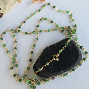 Puede incluir: Un collar de cadena dorada con cuentas de piedras preciosas verdes. El collar está sobre una piedra negra y un trozo de tela.