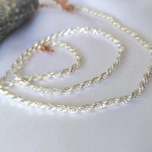 Puede incluir: Una longitud continua de cadena de cuerda de color plateado con una textura metálica trenzada. La cadena está enrollada en un patrón circular suelto, mostrando su superficie reflectante y la estructura uniforme de los eslabones en toda la longitud.