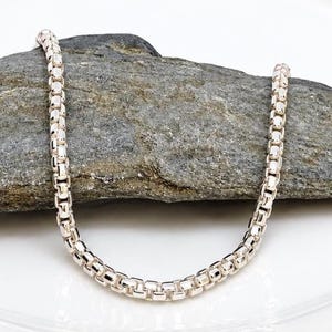 May include: A silver-toned chain necklace featuring a rounded box link design. The chain is composed of small, uniform, square-shaped metallic links connected in a continuous, flexible strand that creates a smooth, polished, and reflective surface.