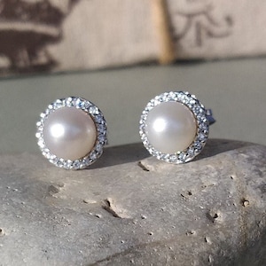 Puede incluir: Un par de pendientes de perlas con un halo de pequeños cristales transparentes. Los pendientes están colocados sobre una piedra gris, con un fondo borroso de una caja de regalo atada con hilo y una cinta azul claro.