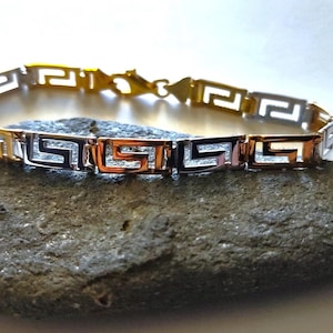 May include: A link bracelet featuring a repeating Greek key pattern. The design alternates between polished gold-tone and silver-tone metal segments, connected by small metal links. The bracelet includes a lobster claw clasp closure.