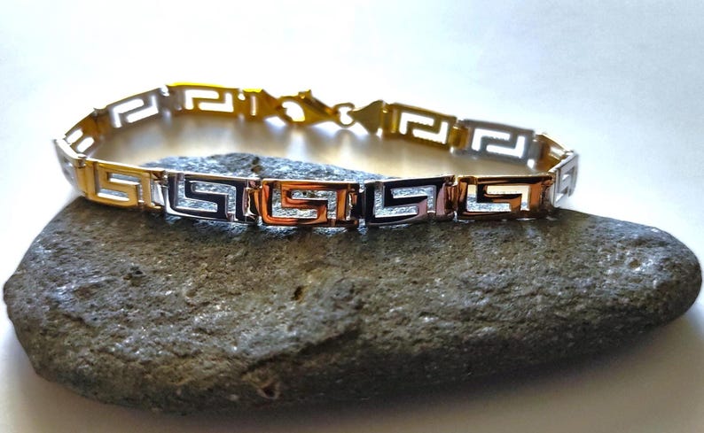 May include: A link bracelet featuring a repeating Greek key pattern. The design alternates between polished gold-tone and silver-tone metal segments, connected by small metal links. The bracelet includes a lobster claw clasp closure.