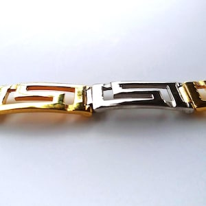 May include: A link bracelet featuring a repeating Greek key pattern, constructed from alternating polished silver-tone and gold-tone metal segments. The design consists of interconnected geometric shapes that form a continuous, structured band.