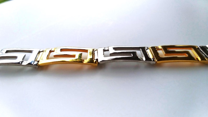 May include: A link bracelet featuring a repeating Greek key pattern, constructed from alternating polished silver-tone and gold-tone metal segments. The design consists of interconnected geometric shapes that form a continuous, structured band.