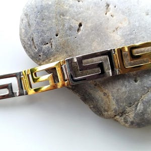 May include: A two-tone bracelet featuring alternating gold-tone and silver-tone rectangular links, each engraved with a repeating Greek key pattern. The metal links are connected in a continuous chain design with a polished, reflective finish.