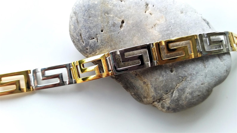 May include: A two-tone bracelet featuring alternating gold-tone and silver-tone rectangular links, each engraved with a repeating Greek key pattern. The metal links are connected in a continuous chain design with a polished, reflective finish.
