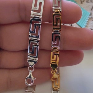 May include: Two link bracelets featuring a repeating Greek key pattern. One bracelet is finished in a polished silver tone, while the other features a two-tone design with alternating silver and gold-colored rectangular links and a lobster claw clasp.