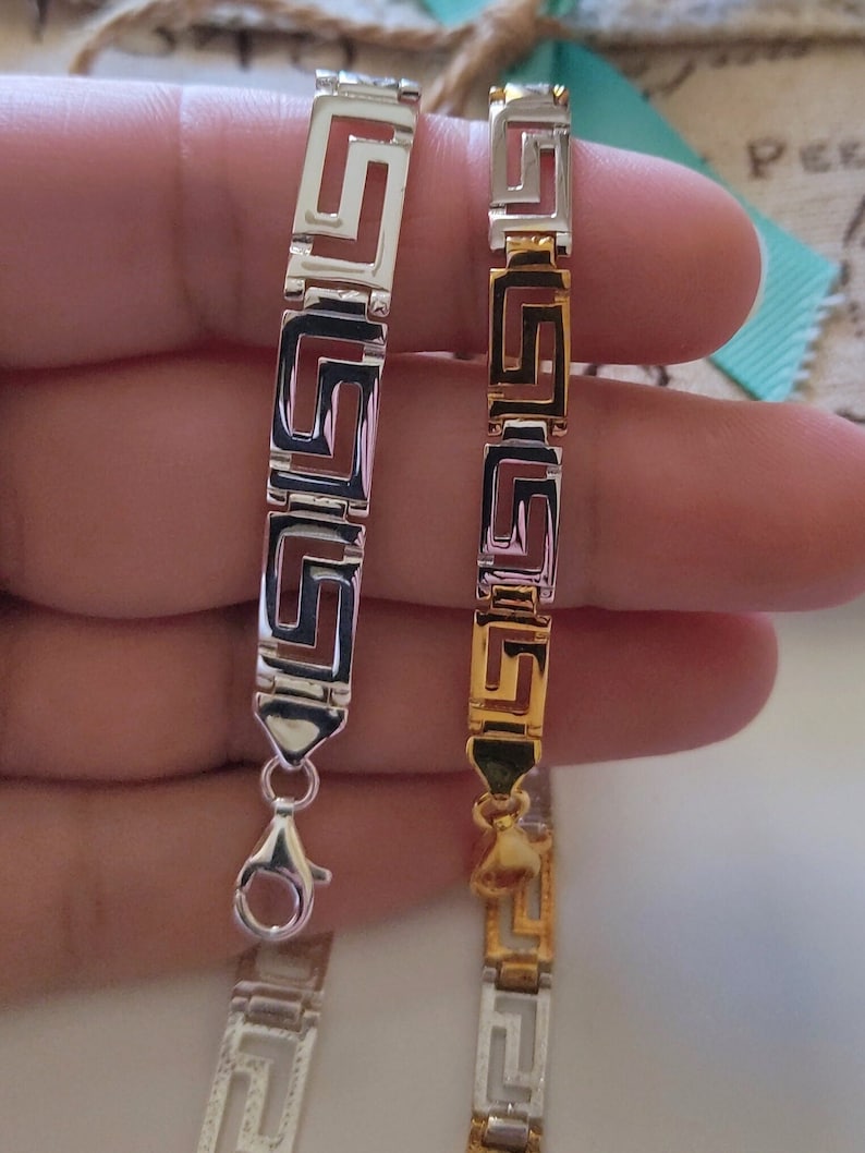 May include: Two link bracelets featuring a repeating Greek key pattern. One bracelet is finished in a polished silver tone, while the other features a two-tone design with alternating silver and gold-colored rectangular links and a lobster claw clasp.