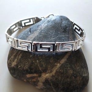 May include: A silver-toned link bracelet featuring a repeating Greek key pattern. The bracelet is constructed from multiple rectangular segments, each containing a cut-out geometric design, connected by small hinges to form a continuous circular band.