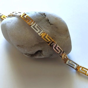 May include: A two-tone bracelet featuring a repeating Greek key pattern. The design alternates between polished gold-tone and silver-tone rectangular links, connected to form a continuous chain. A small gold-tone lobster claw clasp is at the end.