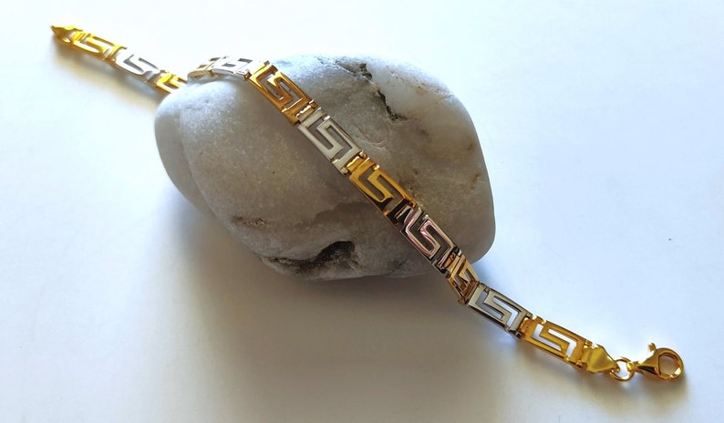 May include: A two-tone bracelet featuring a repeating Greek key pattern. The design alternates between polished gold-tone and silver-tone rectangular links, connected to form a continuous chain. A small gold-tone lobster claw clasp is at the end.