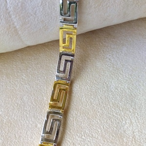 May include: A link bracelet featuring alternating gold-tone and silver-tone rectangular segments. Each segment displays a cut-out Greek key pattern. The bracelet is constructed from a series of these repeating geometric metal links in a straight line.