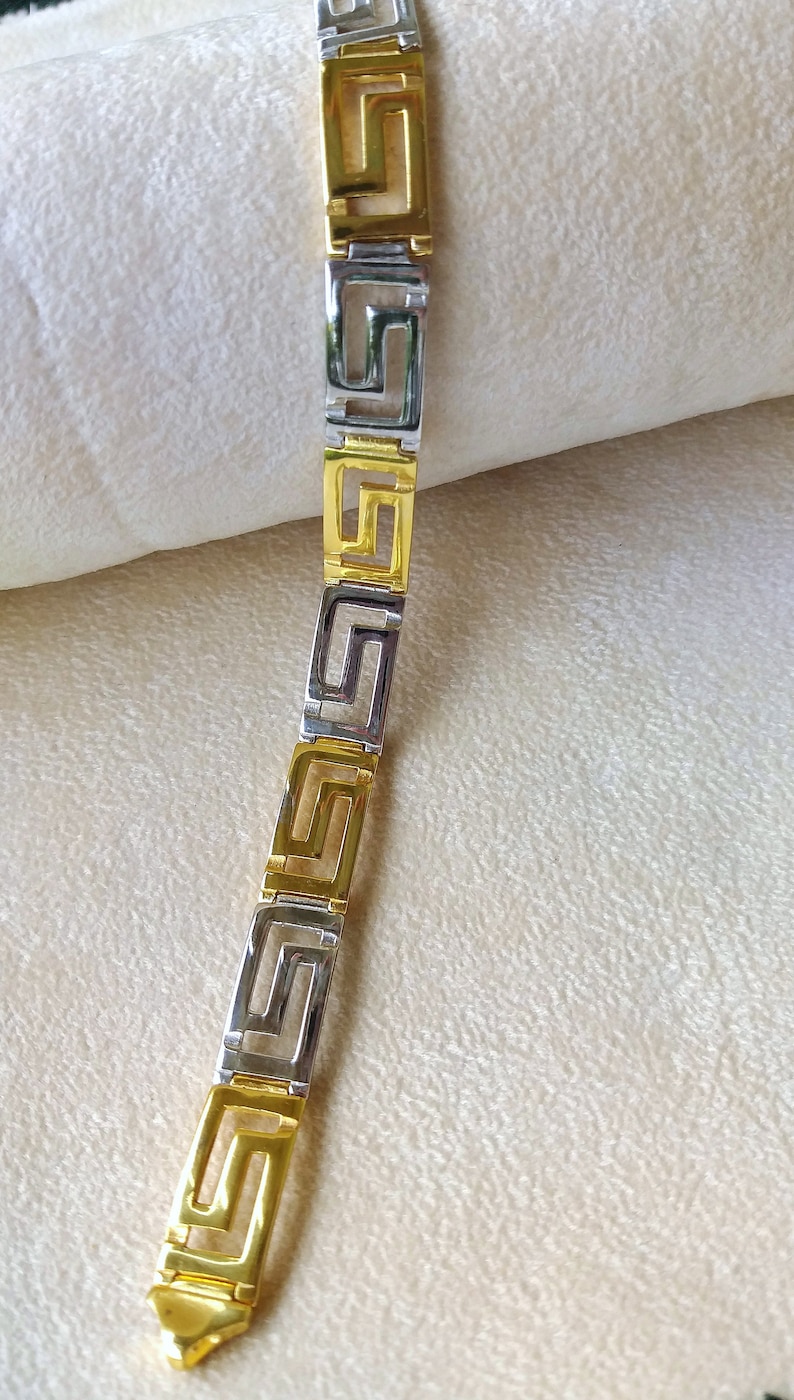 May include: A link bracelet featuring alternating gold-tone and silver-tone rectangular segments. Each segment displays a cut-out Greek key pattern. The bracelet is constructed from a series of these repeating geometric metal links in a straight line.