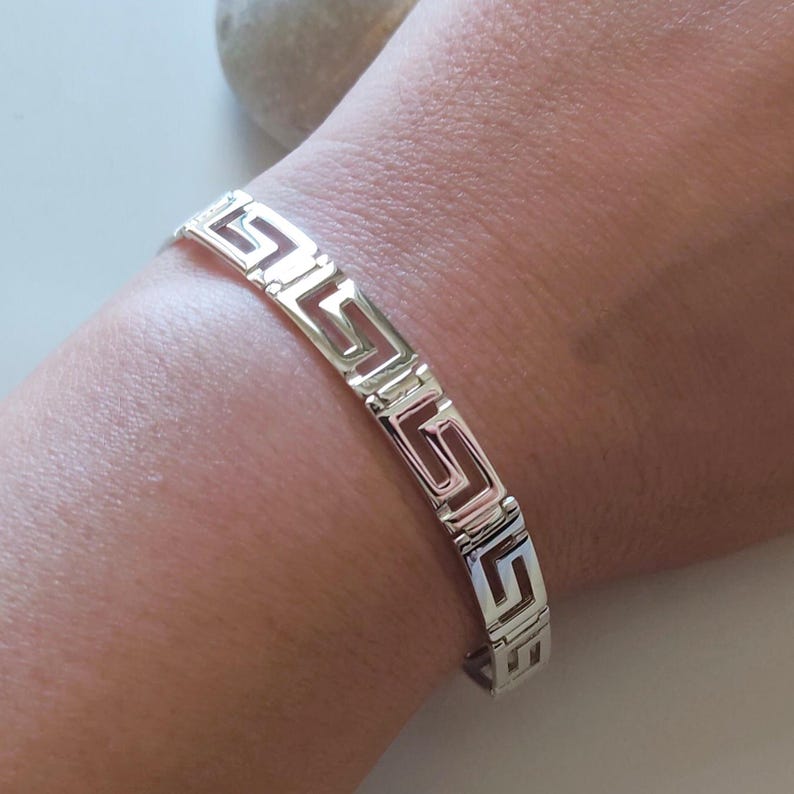 May include: A silver-toned metal bracelet featuring a repeating Greek key pattern. The bracelet is constructed from a series of interconnected, angular geometric links that form a continuous, open-work design around the wrist, showcasing a polished finish.