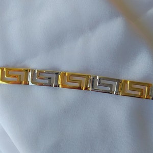 May include: A link bracelet featuring alternating gold and silver-toned rectangular segments. Each segment displays a cut-out Greek key pattern, creating a repeating geometric design across the entire length of the metal jewelry piece.