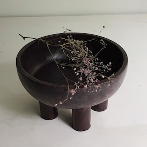 May include: A dark brown wooden bowl with three legs, sitting on a white surface. Dried flowers are inside the bowl.