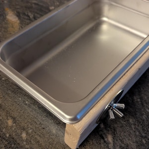 May include: A stainless steel rectangular container set in a wooden frame, secured with wing nuts. The metal tray is shiny and reflective. The wooden frame has a light color. The item is on a speckled countertop.