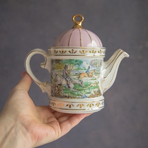 Könnte beinhalten: Vintage Porzellan-Teekanne mit rosa Deckel und goldenen Akzenten. Die Teekanne zeigt eine Jagdszene mit Reitern auf Pferden. Sie hat einen weißen Korpus, goldene Verzierungen und wird in einer Hand gehalten.