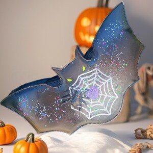 Op de afbeelding: Een vleermuisvormige handtas in grijstinten en blauw, versierd met een wit spinnenweb en een paarse hart. Een zwarte spin zit op het web. De tas is besprenkeld met kleurrijke glitter. Kleine oranje pompoenen en een uitgesneden pompoen staan op de achtergrond.