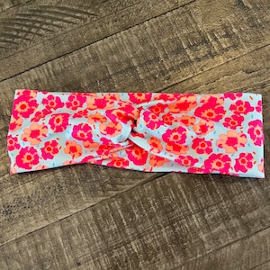May include: A floral headband featuring a light blue background with a pattern of pink and orange flowers. The headband is twisted at the center, creating a stylish accessory. The fabric appears soft and comfortable, suitable for various occasions.