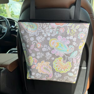 May include: A hanging car trash bag featuring a gray fabric exterior with a colorful paisley and floral pattern in shades of pink, yellow, and teal. The bag is suspended by two black straps attached to the headrest of a vehicle seat.
