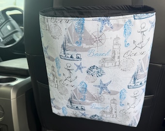 May include: A hanging car trash bag featuring a nautical print on a white background with blue and gray illustrations of sailboats, lighthouses, anchors, seahorses, starfish, and compasses, with the words Ocean and Beach printed throughout.