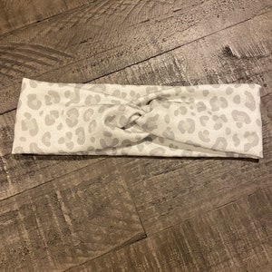 May include: A white headband with a gray leopard print pattern. The fabric is twisted at the center. The headband is designed to be worn around the head.