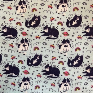 May include: Light blue fabric with a repeating pattern of black and white cats, planets, UFOs, rainbows, clouds, and stars. The cats are in various poses. The design has a whimsical, space-themed aesthetic.