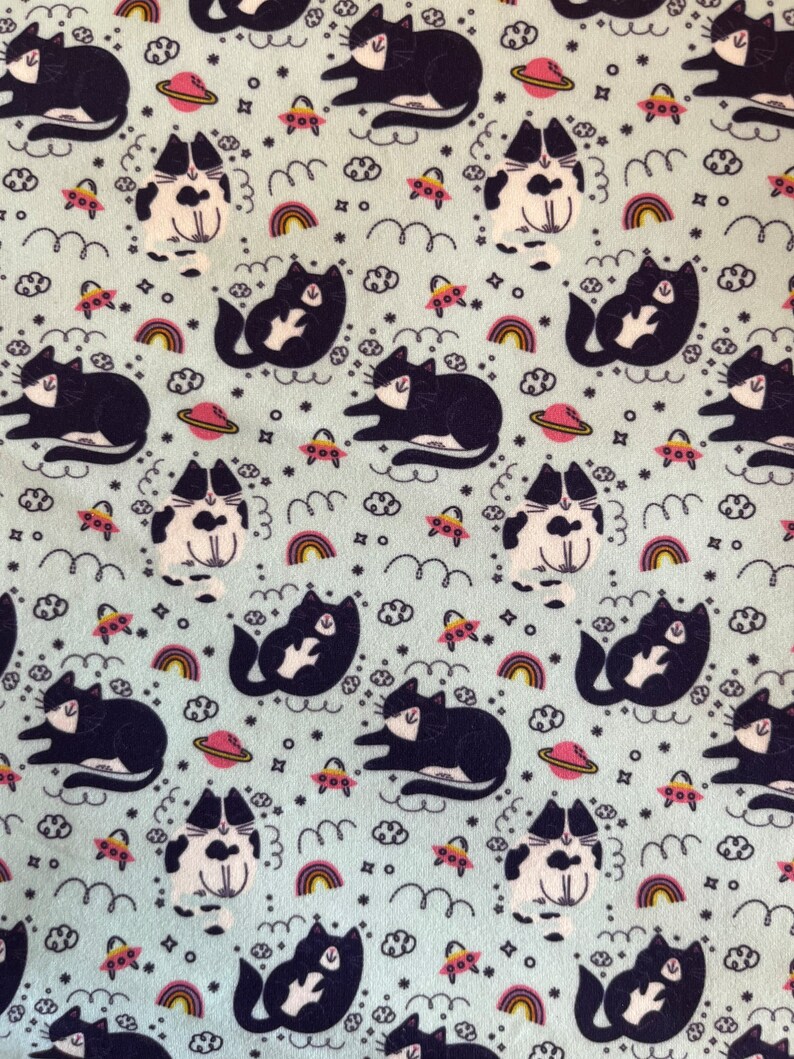 May include: Light blue fabric with a repeating pattern of black and white cats, planets, UFOs, rainbows, clouds, and stars. The cats are in various poses. The design has a whimsical, space-themed aesthetic.