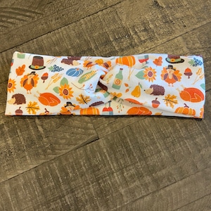 May include: White headband with a Thanksgiving-themed pattern. The design includes images of turkeys, pumpkins, sunflowers, and other fall elements. The headband is made of a soft fabric and is twisted at the top.