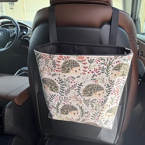 May include: A fabric car trash bag featuring a repeating pattern of hedgehogs, red berries, and green foliage. The bag has a black interior lining and is suspended by two black straps attached to the headrest of a brown leather vehicle seat.