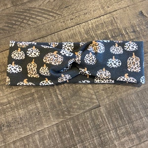 May include: A dark gray headband featuring a repeating pattern of pumpkins with leopard print and white spots. The pumpkins have brown stems. The headband is twisted at the center.