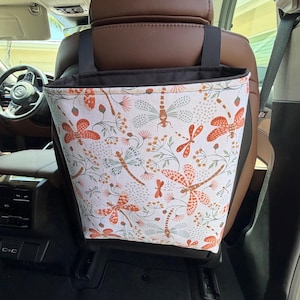 May include: A car trash bag hanging from the back of a vehicle seat, featuring a white fabric exterior with a repeating pattern of orange and sage green dragonflies and botanical elements, finished with a black fabric trim and black hanging straps.