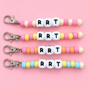 May include: Four beaded keychains featuring white square beads with the black letters R, R, and T. Each keychain has a silver-toned metal lobster clasp and a string of colorful round beads in shades of pink, yellow, orange, and light blue.