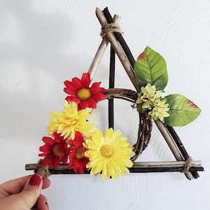 以下が含まれることがあります： 中央に編まれたリースのある木製の三角形。黄色と赤の花と緑の葉で飾られています。